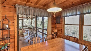 L’intérieur d’un chalet en bois comprend une table à manger, des chaises, un lustre et de grandes fenêtres donnant sur une zone boisée.