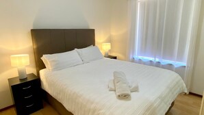 A neatly made bed with white linens and a folded towel, two bedside lamps, and a window with sheer curtains.