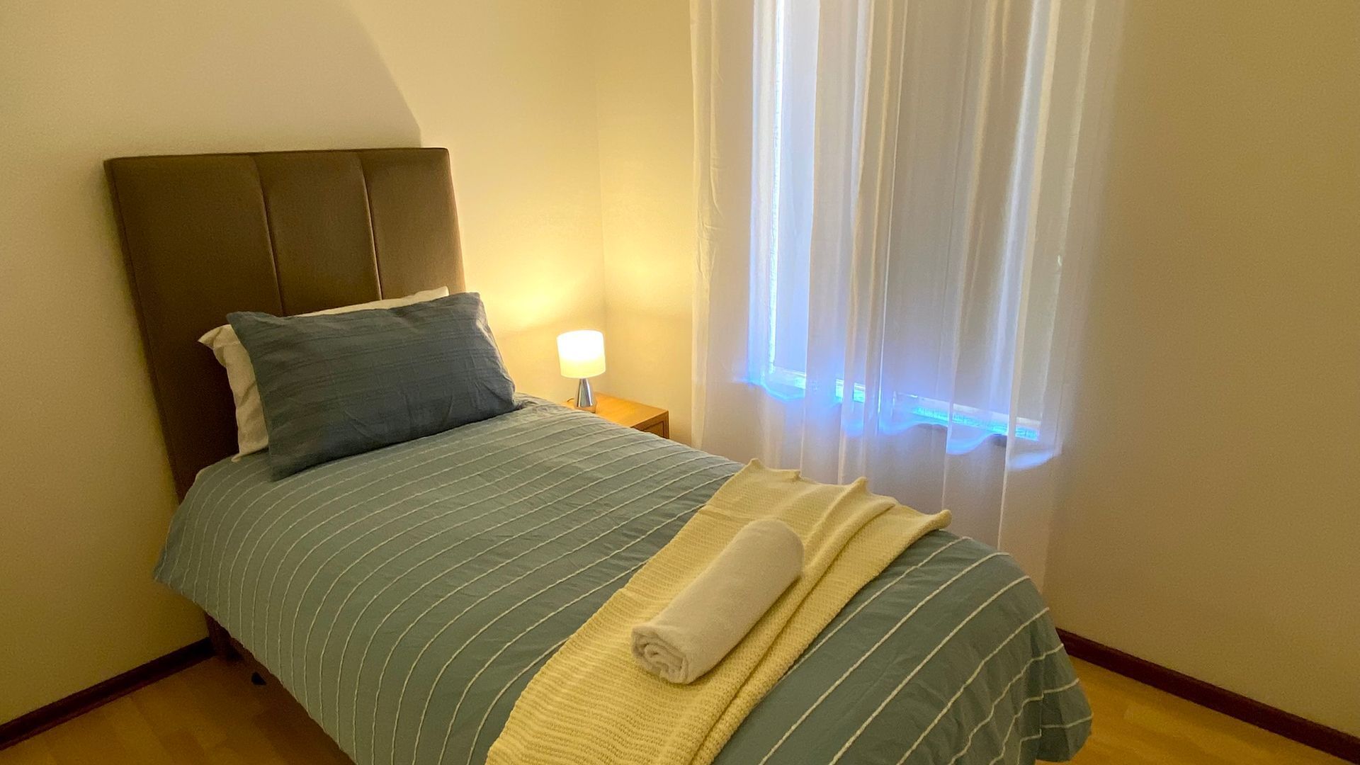 Ein ordentlich bezogenes Bett mit einer gestreiften Tagesdecke, ein zusammengerolltes Handtuch und ein Nachttisch mit Lampe.