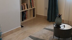 Une pièce avec une bibliothèque, une petite table ronde sur laquelle se trouve un ananas, une fenêtre avec des rideaux et un tapis à motifs.