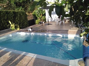 Piscina com deck ladrilhado, rodeada por vegetação e cadeiras brancas.