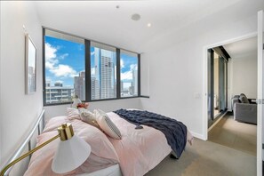A modern bedroom with a large window, a bed with a striped bedspread, a bedside lamp, and a view of the city skyline. - Comfy 2BR Apt Near Southern Cross with City Views (Melbourne)
