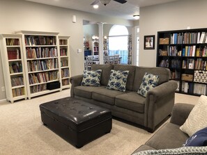 Un salon avec un canapé gris, un pouf noir et deux bibliothèques blanches remplies de livres.