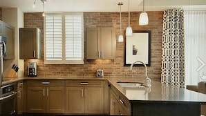 A modern kitchen with wooden cabinets, a built-in oven, a coffee maker, pendant lights, and a large island with a sink.