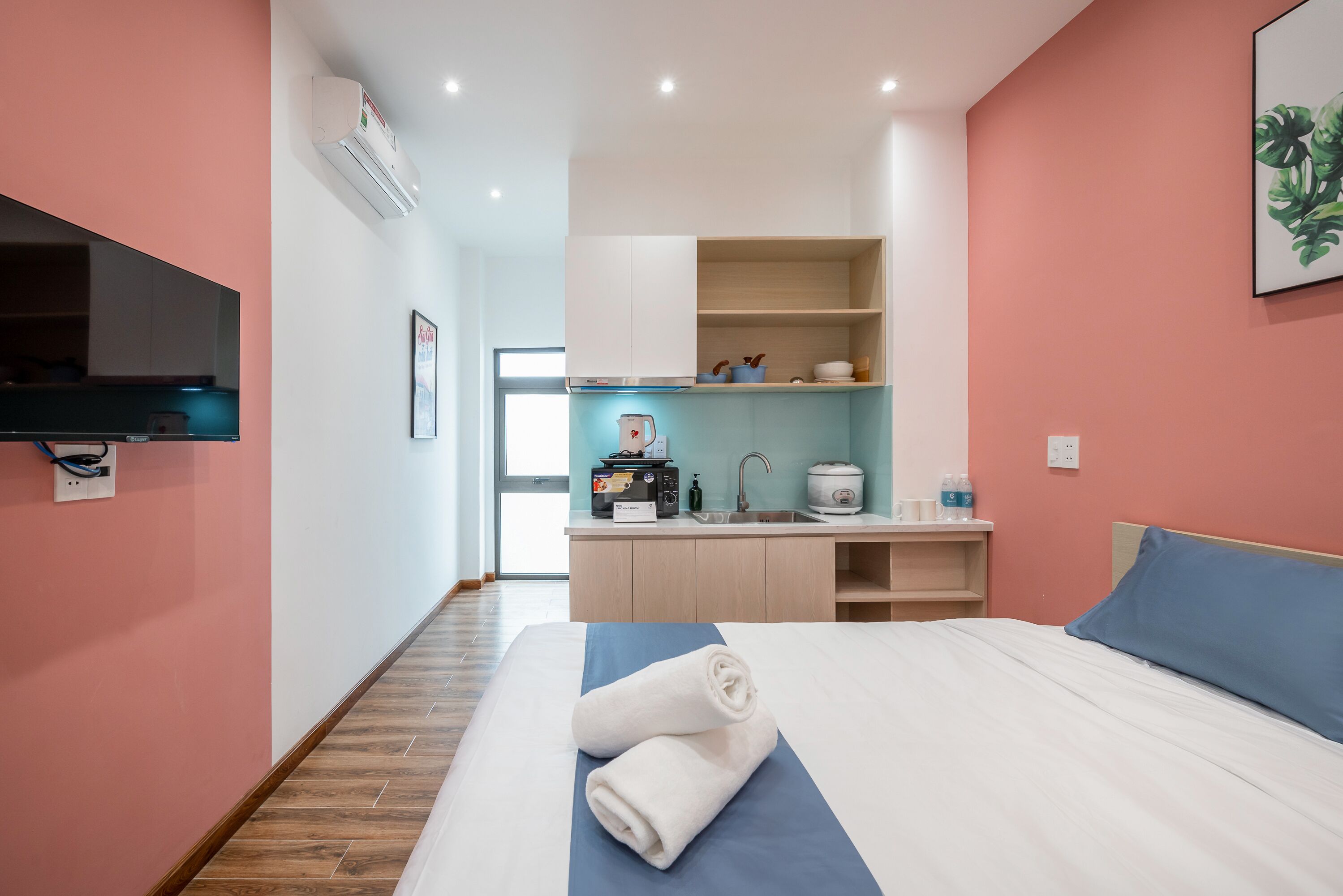 A hotel room with a bed, a TV mounted on the wall, a kitchenette, and a bathroom visible through an open door.