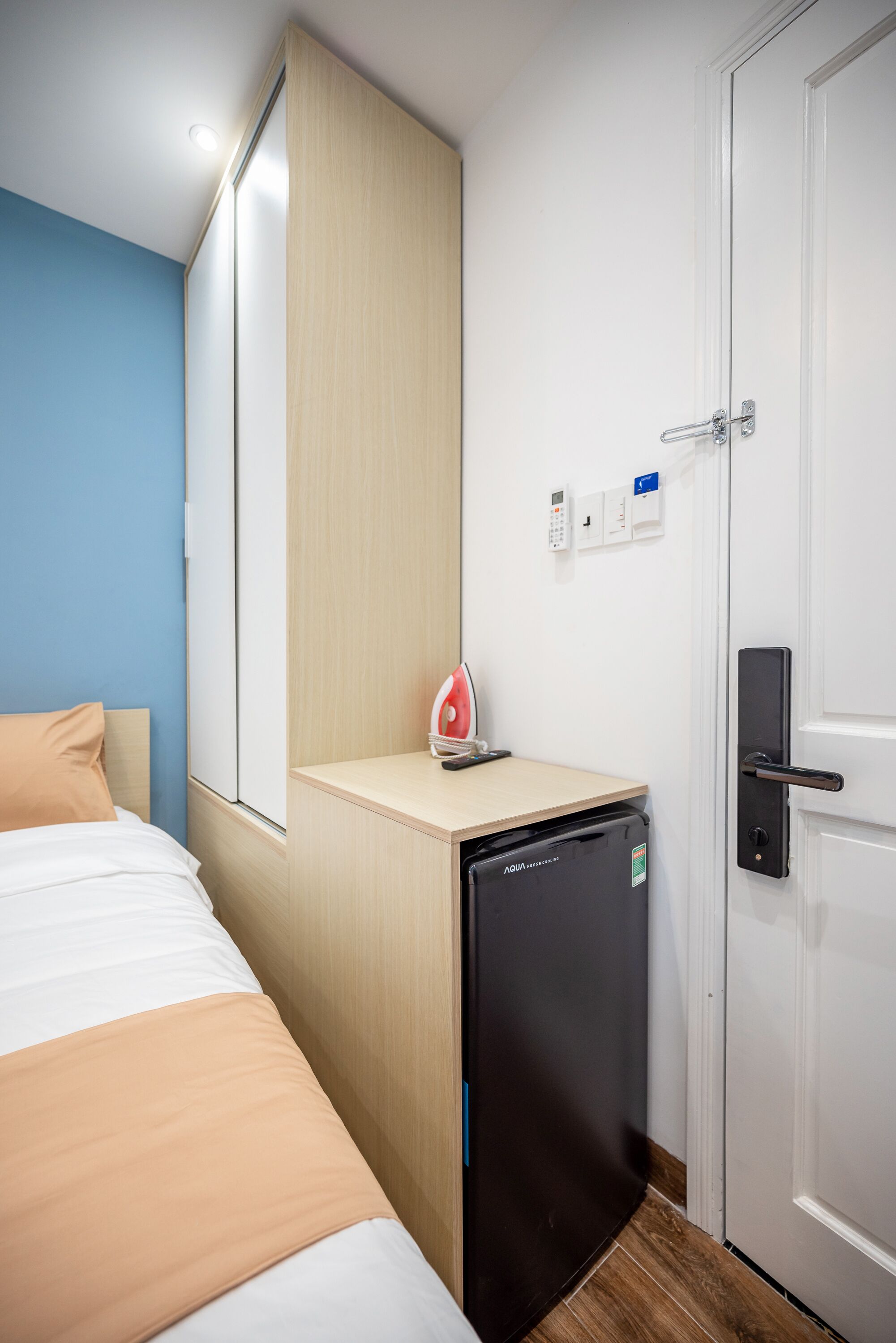 A hotel room with a bed, a small desk with a water dispenser, a white door, and a light switch on the wall.