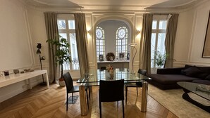 Een ruime woonkamer met een glazen eettafel, een bank en een salontafel. Er zijn grote ramen met gordijnen, een radiator en een potplant.
