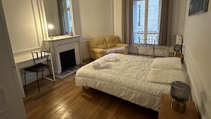 Een slaapkamer met een groot bed, een stoel, een open haard en een raam met gordijnen.