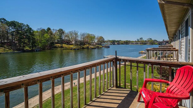 Una terraza de madera con baranda que da a un lago tranquilo, una silla Adirondack roja y un edificio con porche cubierto.
