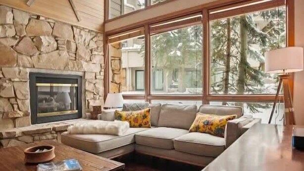 A cozy living room with a stone fireplace, a large window with a view of trees, a sectional sofa, and a coffee table.