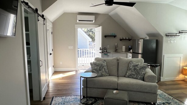 A spacious living room with a gray sofa, ottoman, and patterned rug. A ceiling fan and an air conditioning unit are visible.