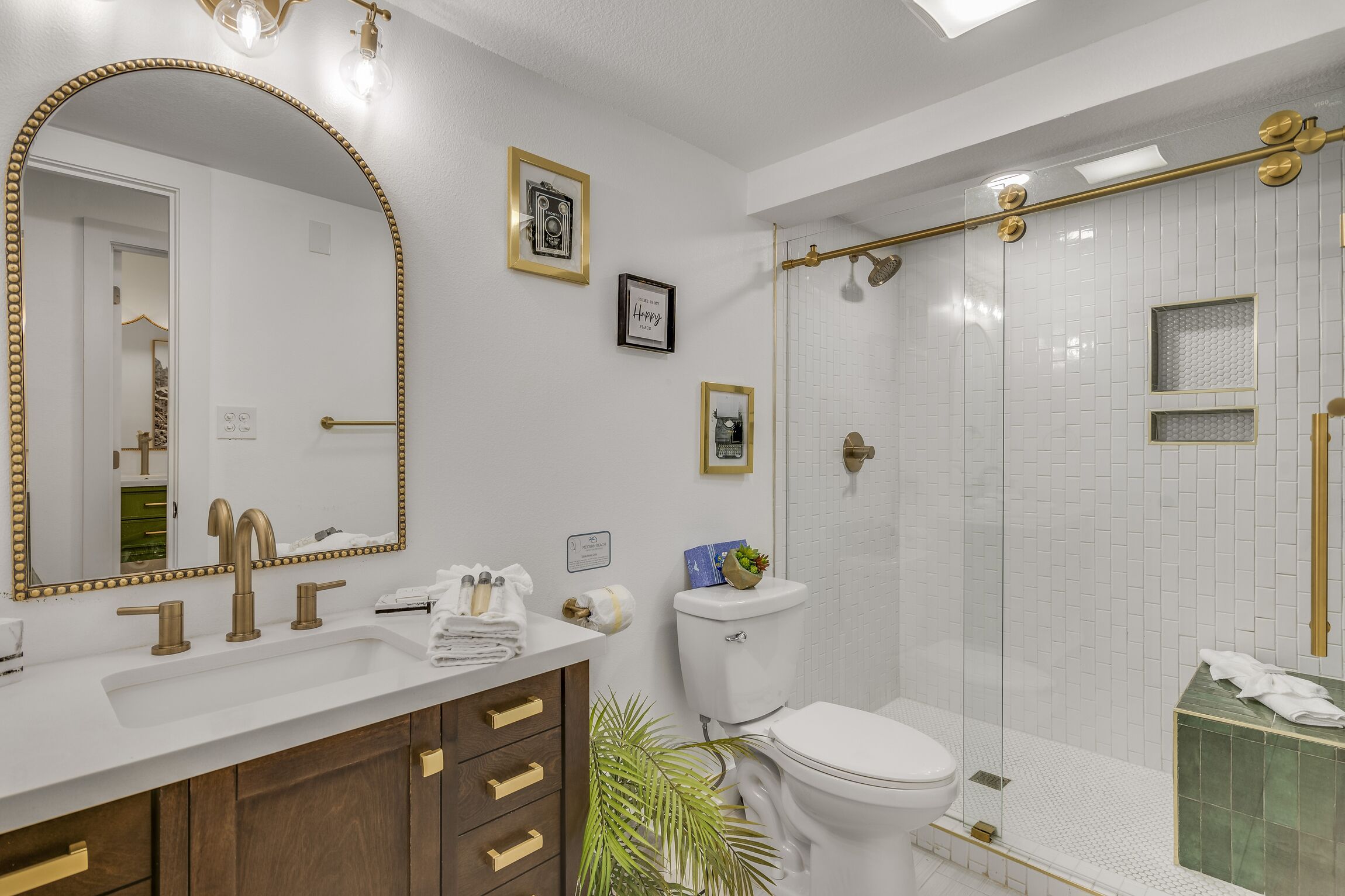 Ein Badezimmer mit Waschbecken in Weiß, goldenen Armaturen, einem großen Spiegel, einer Toilette und einer Duschkabine aus Glas.