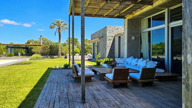 A modern outdoor patio with wooden furniture, a stone wall, and a glass door leading to a room. - Luxury 10,000 sq ft modern house with breathtaking ocean views and every amenity (José Ignacio)