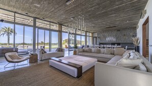 A spacious living area with a large sofa, a coffee table, and a chair. There is a stone fireplace and a view of the outdoors through large windows. - Luxury 10,000 sq ft modern house with breathtaking ocean views and every amenity (José Ignacio)