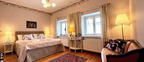 A hotel room with a bed, bedside tables, a chair, a window with curtains, a radiator, and a chandelier.