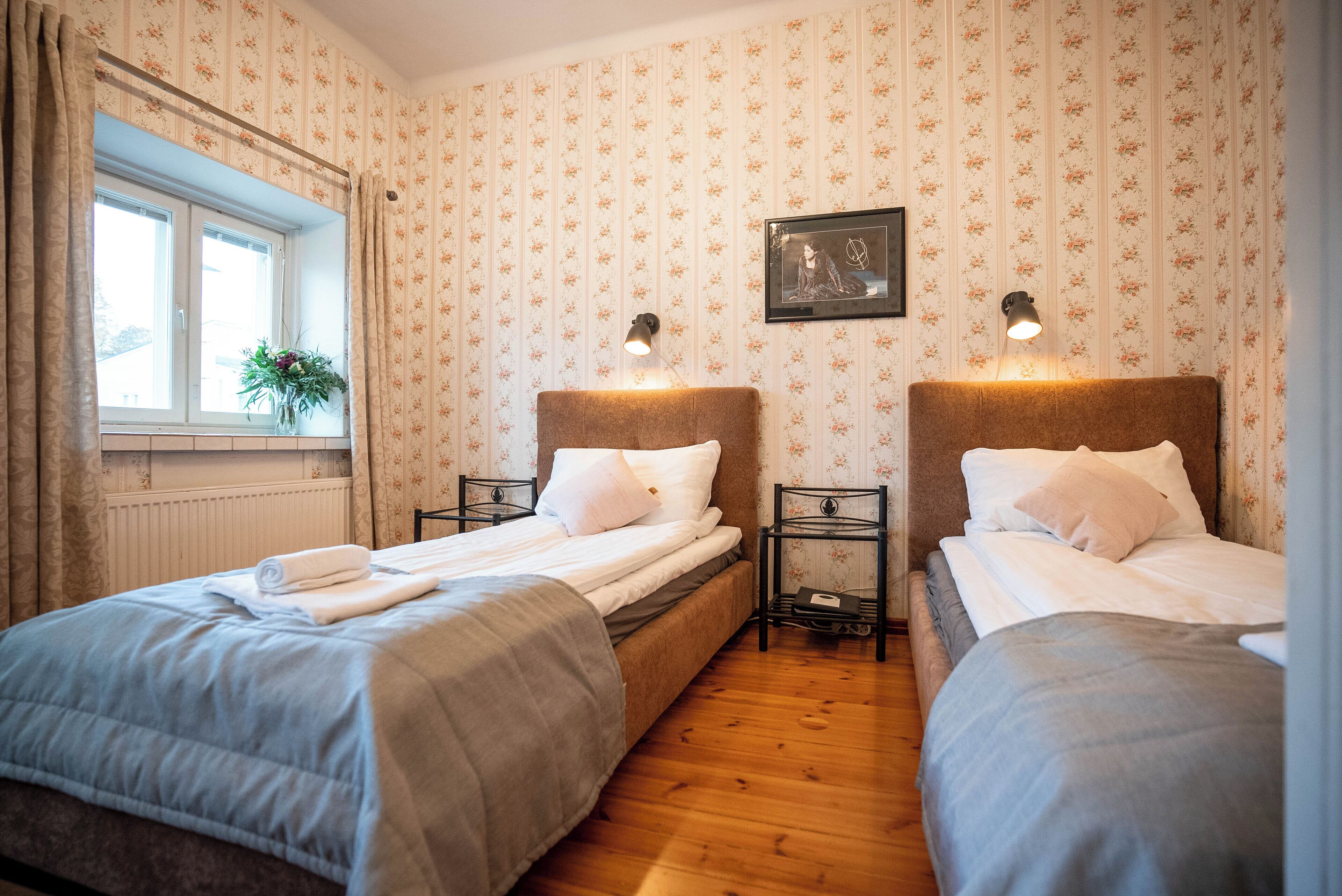 Un dormitorio con dos camas individuales, una ventana con cortinas, un radiador y un cuadro enmarcado en la pared.
