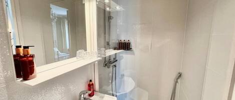 Une salle de bain avec un lavabo blanc, un miroir et une douche. On peut voir des articles de toilette sur l’étagère.
