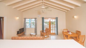 A spacious room with a high ceiling, a ceiling fan, a dining table with chairs, a sofa, a TV on a stand, and a window with curtains. - Neem View Apartments Antigua (Falmouth Harbour)