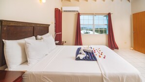 A hotel room with a large bed, a wooden headboard, a window with curtains, an air conditioning unit, and a wooden door. - Neem View Apartments Antigua (Falmouth Harbour)