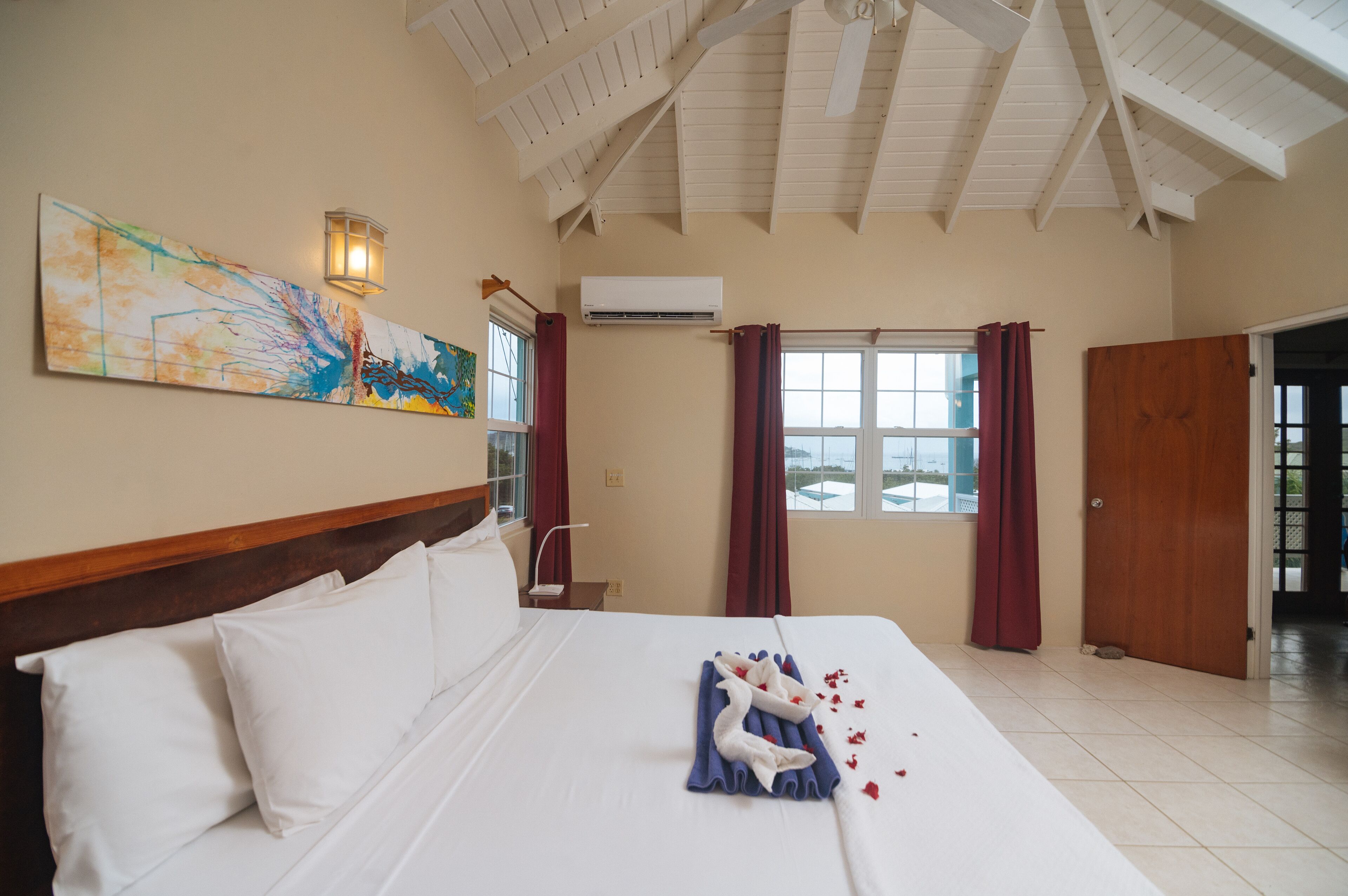 Une chambre d’hôtel avec un lit, un tableau, un appareil de climatisation, une fenêtre avec des rideaux et une porte en bois.