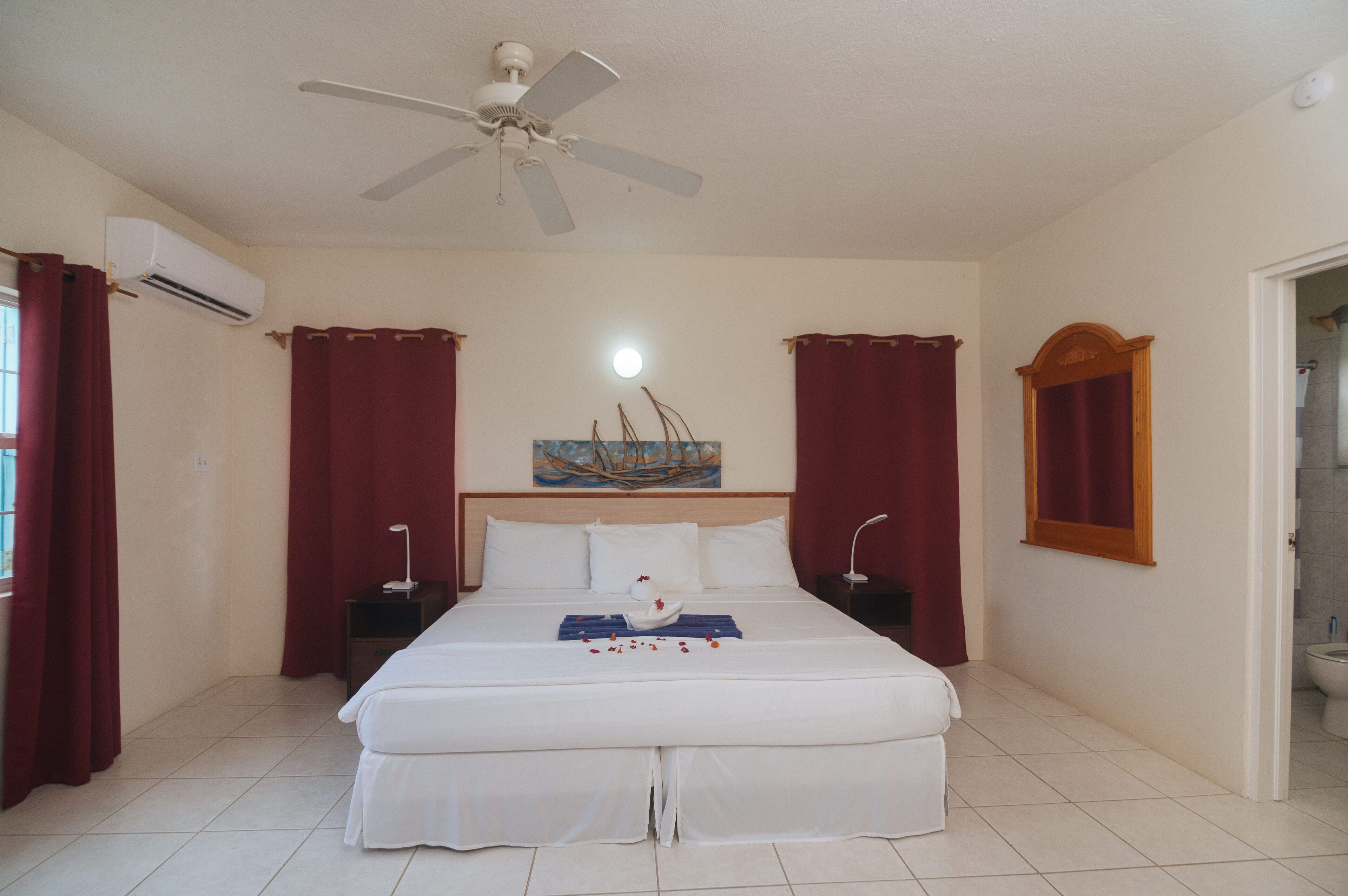 A hotel room with a bed, a ceiling fan, an air conditioning unit, a painting on the wall, and a window with curtains.