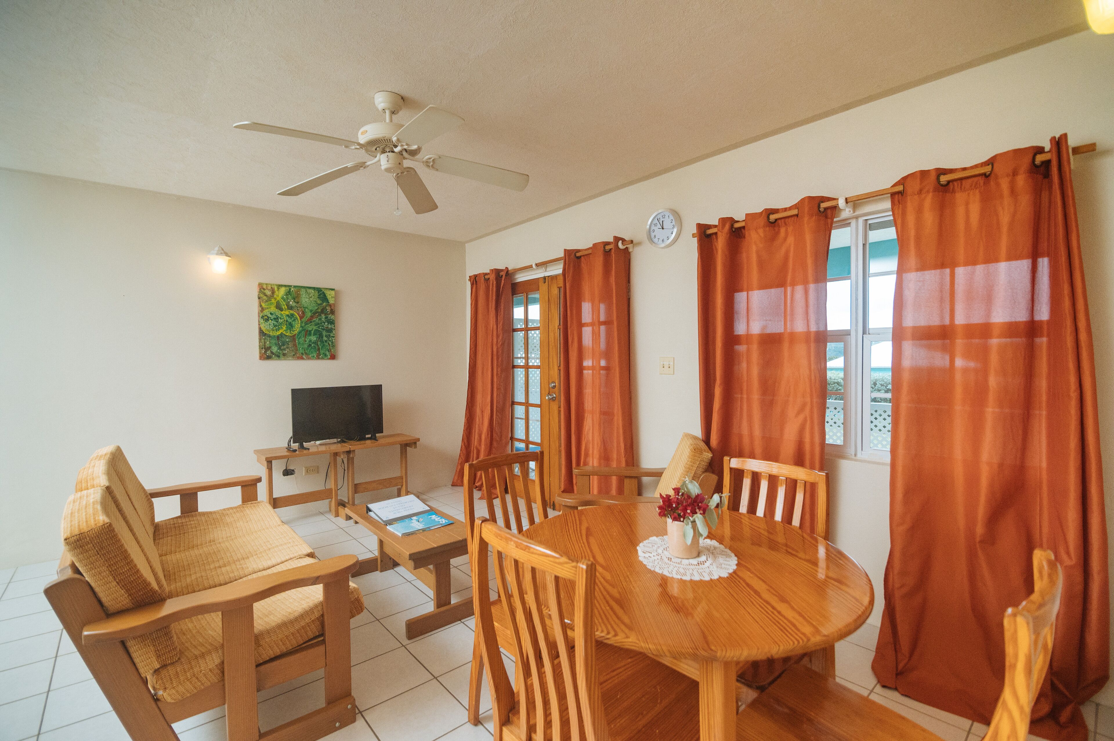 A dining area with a wooden table and chairs, a ceiling fan, a television on a stand, and orange curtains.