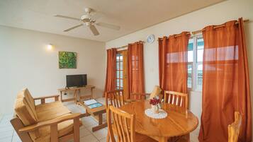 A dining area with a wooden table and chairs, a ceiling fan, a television on a stand, and orange curtains.