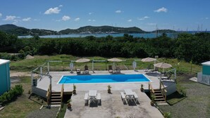 A swimming pool with lounge chairs and umbrellas, surrounded by greenery and a view of the sea and hills. - Neem View Apartments Antigua (Falmouth Harbour)