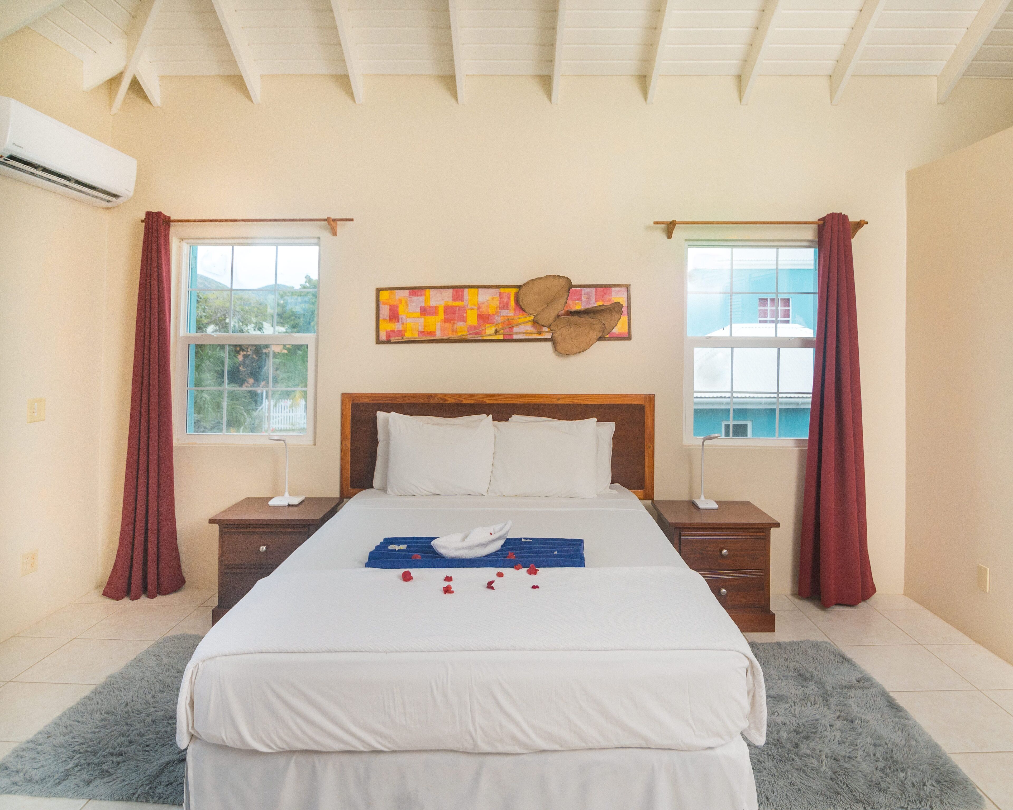 Quarto de hotel com uma cama grande, duas mesas de cabeceira, uma janela com cortinas e um ventilador de teto.