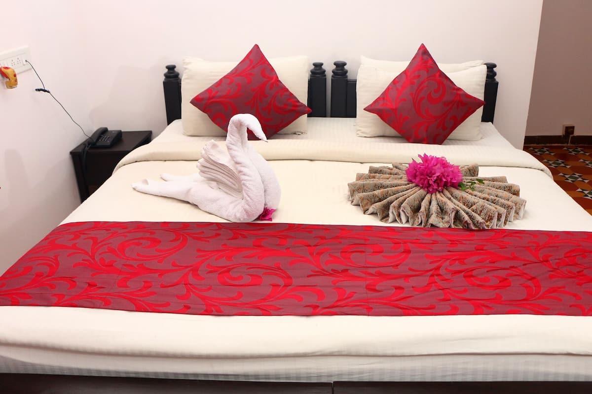 A bed with a red and white patterned bedspread, two red pillows, a white towel, and a decorative floral arrangement.