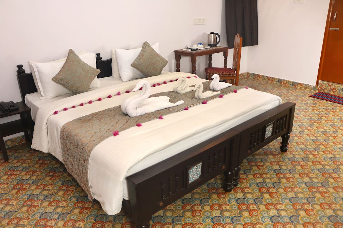 A neatly made bed with white linens and decorative swan pillows, a bedside table with a kettle, and a patterned carpet.
