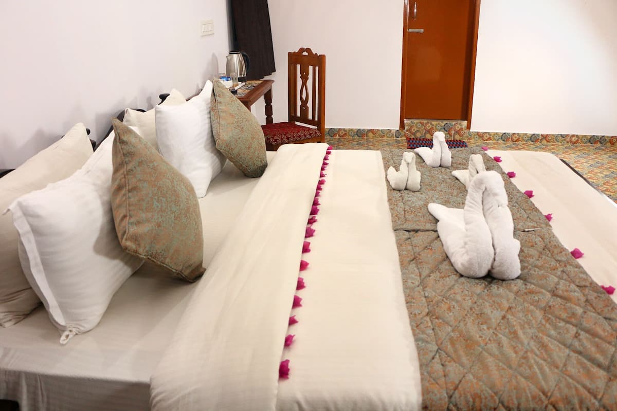 A hotel room with a bed, pillows, a wooden chair, a small table with a kettle, and a door.