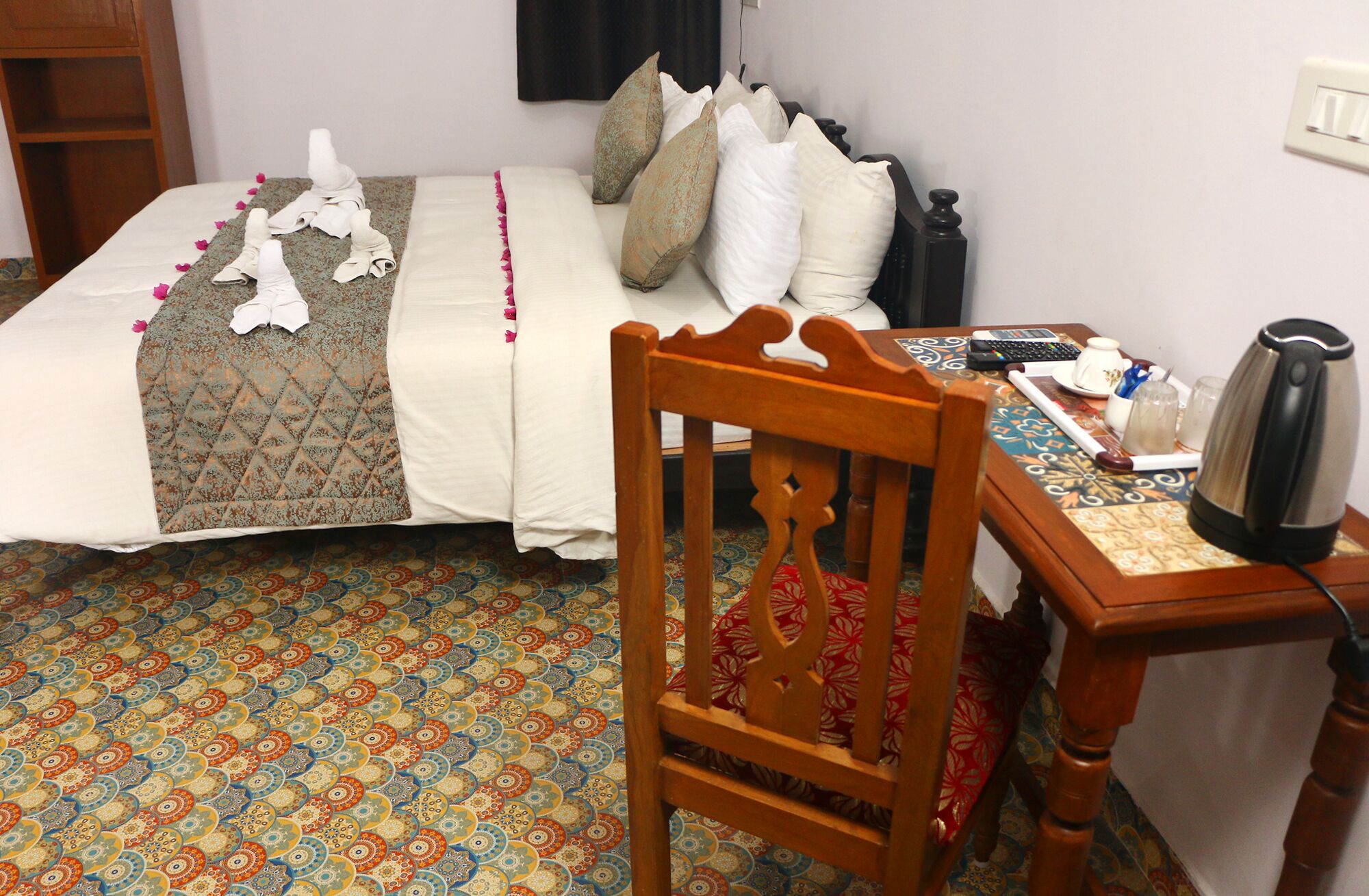 A neatly made bed with a patterned bedspread, a wooden chair, a small table with a kettle, and a patterned carpet.