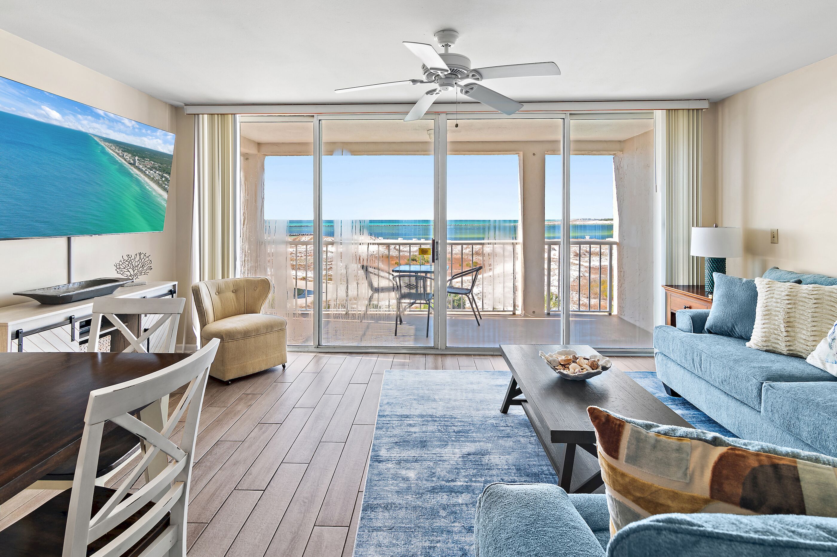 A spacious living room with a large TV, a dining area, and a balcony with a view of the beach.
