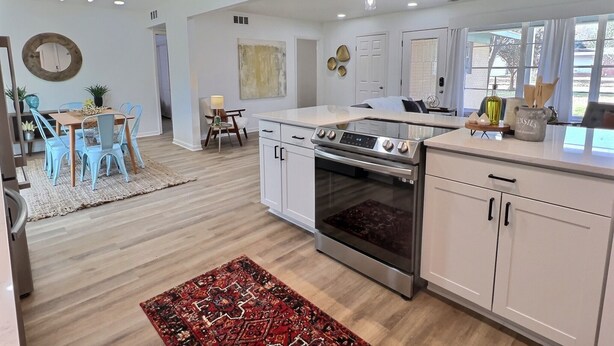 A modern kitchen with white cabinetry, a built-in oven, and a dining area with blue chairs and a wooden table. - Remodeled home with firepit! 1 mile to Randolph AFB & 3 parks! (Universal City)