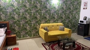 Une pièce avec un canapé jaune, une table basse en verre, un papier peint à motifs, un lit avec un tapis rouge et un petit meuble.