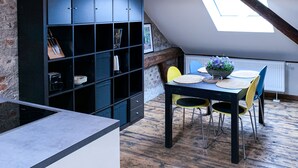 Un espace repas doté d’un plafond haut, de poutres apparentes en bois et d’une lucarne. On y trouve une bibliothèque noire et une table avec des chaises jaunes.