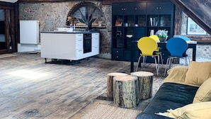 Un espace de vie aménagé dans les combles, avec des poutres apparentes, un coin cuisine et un espace salon comprenant un canapé en cuir noir et des fauteuils colorés.