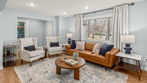 A living room with a beige sofa, two armchairs, a coffee table, and a window with curtains. - Spacious Family Haven - Walk to Downtown & JBU (Siloam Springs)