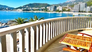 Un balcon avec une table et des chaises en bois, donnant sur une zone côtière avec des bâtiments et des montagnes en arrière-plan.