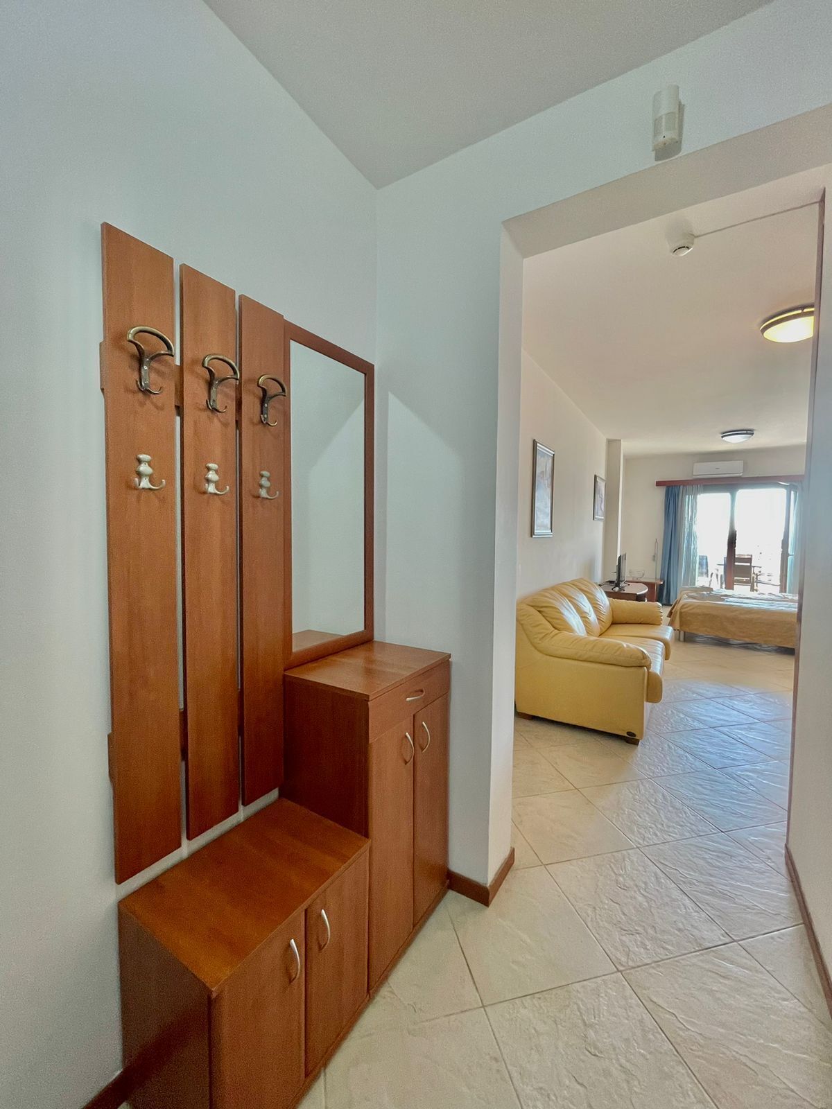 A hotel room with a wooden wardrobe, a mirror, a beige sofa, and a view of the living area.