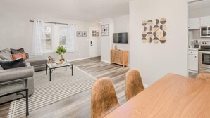 En moderne stue med sofa, sofabord og spisebord. Et fjernsyn er monteret på væggen, og et køkkenområde kan ses i baggrunden.