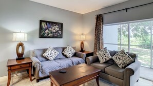 A living room with a sofa, a coffee table, and two side tables with lamps. There is a painting on the wall and a window with curtains. - Super close to Disney, 4BR/3BA,Next to the mall(restaurants) (Kissimmee)