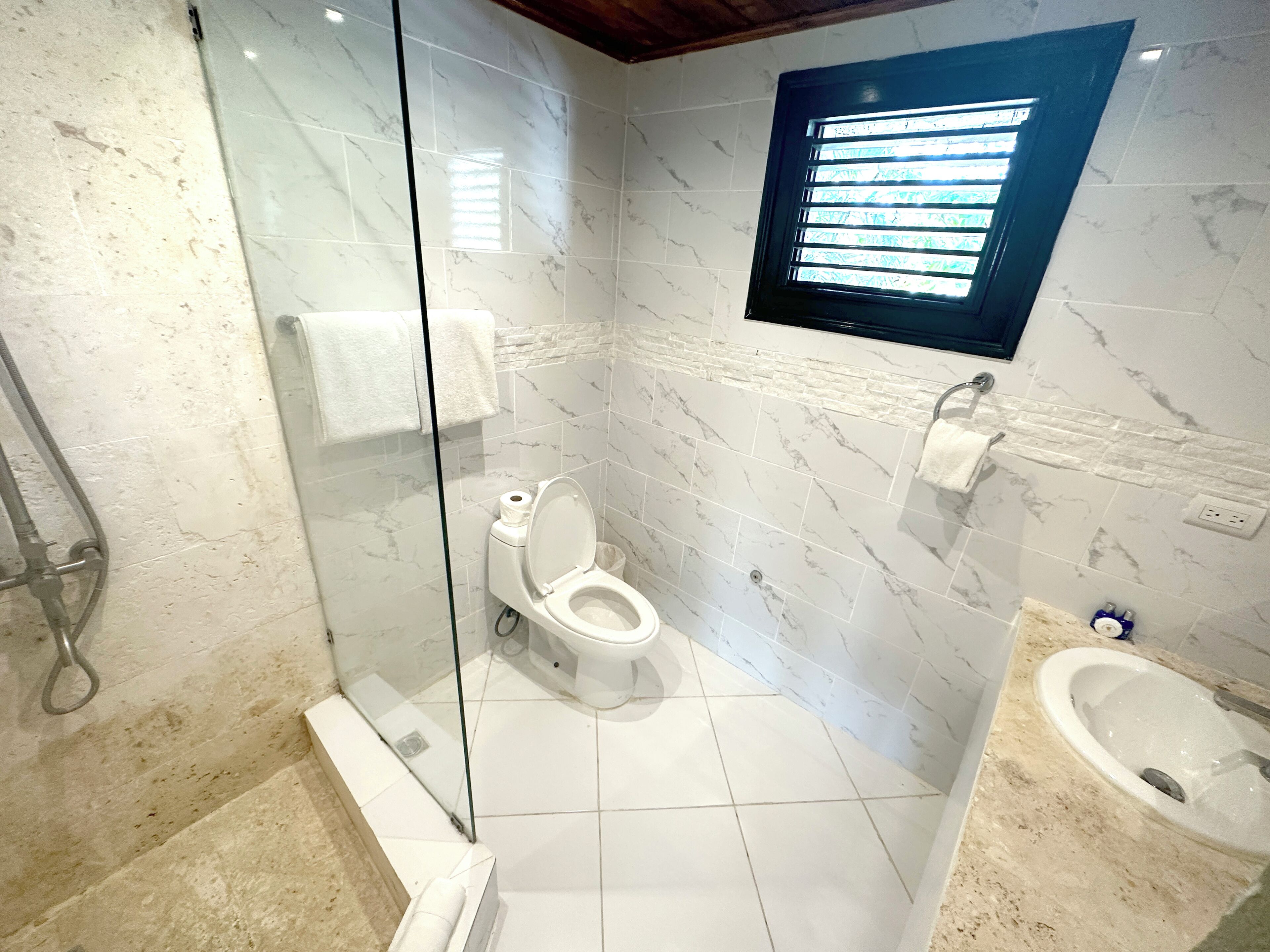 Un baño moderno con ducha de vidrio, inodoro, lavamanos y ventana con persianas.