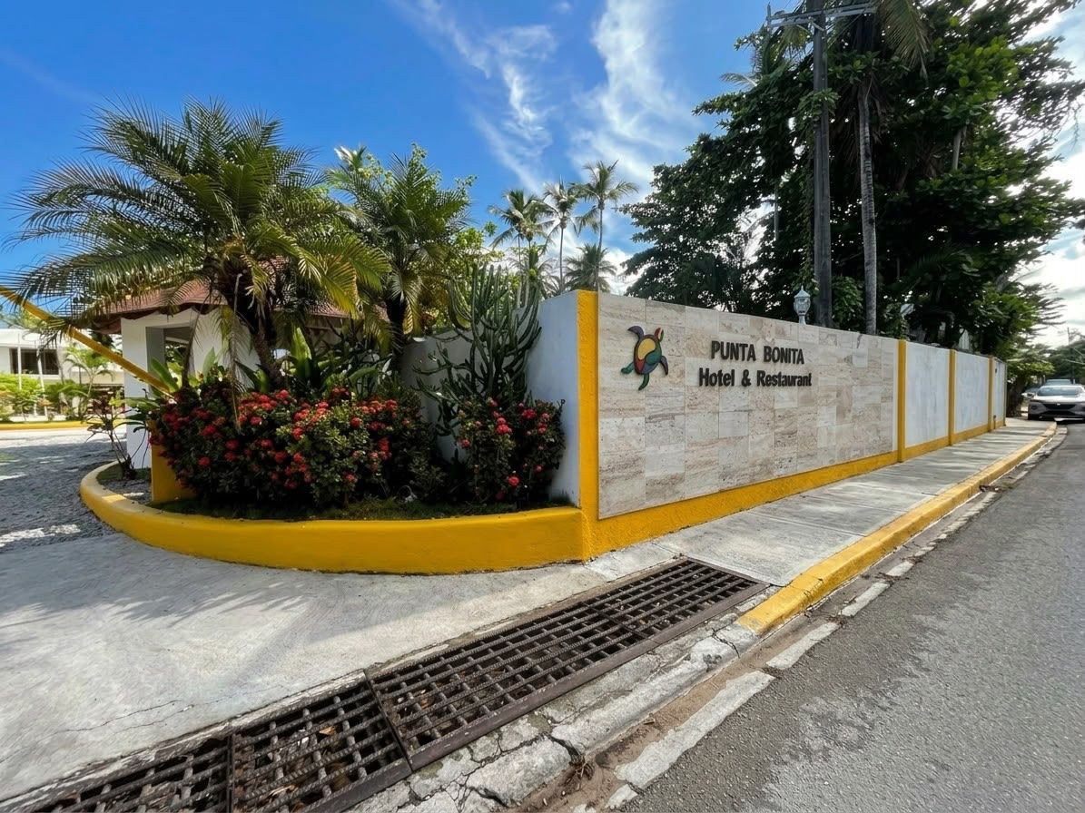 A hotel entrance with a sign for Punta Bonita Hotel & Restaurant, surrounded by tropical plants and a clear sky.