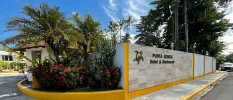 A hotel entrance with a sign for Punta Bonita Hotel & Restaurant, surrounded by tropical plants and a clear sky.