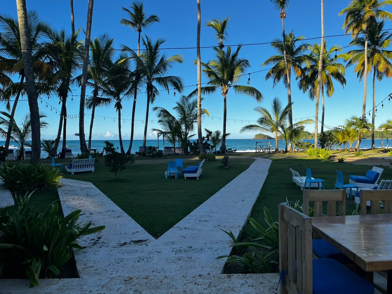 Ein tropisches Resort mit einem Weg zu einer Sitzgelegenheit mit Stühlen und Tisch, umgeben von Palmen und Blick auf den Ozean.