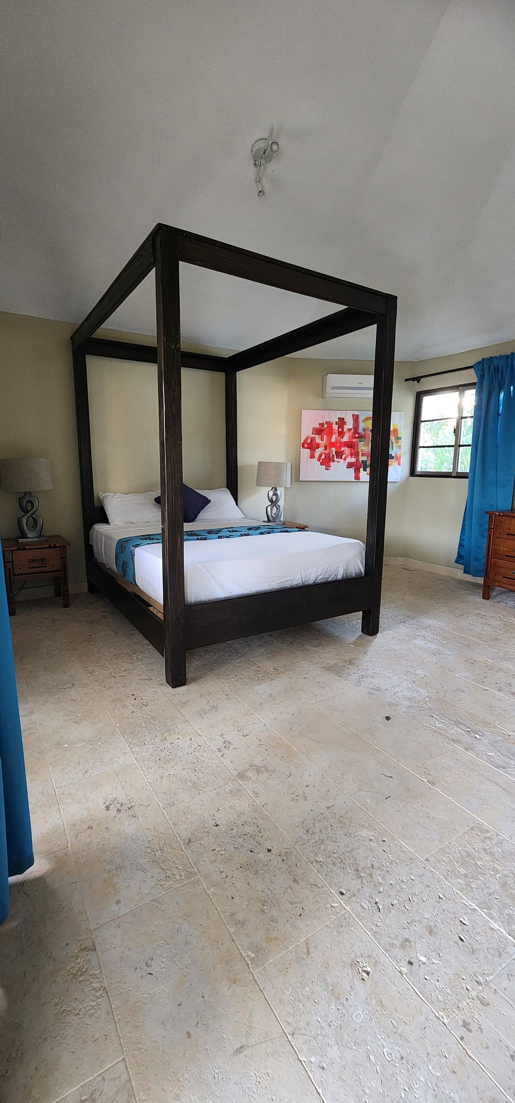 Una cama con dosel en una habitación con piso de baldosas, una mesita de noche con lámpara y una ventana con cortinas.
