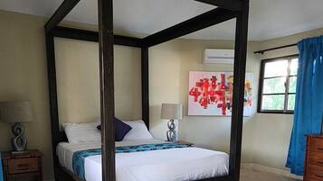 Una cama con dosel en una habitación con piso de baldosas, una mesita de noche con lámpara y una ventana con cortinas.