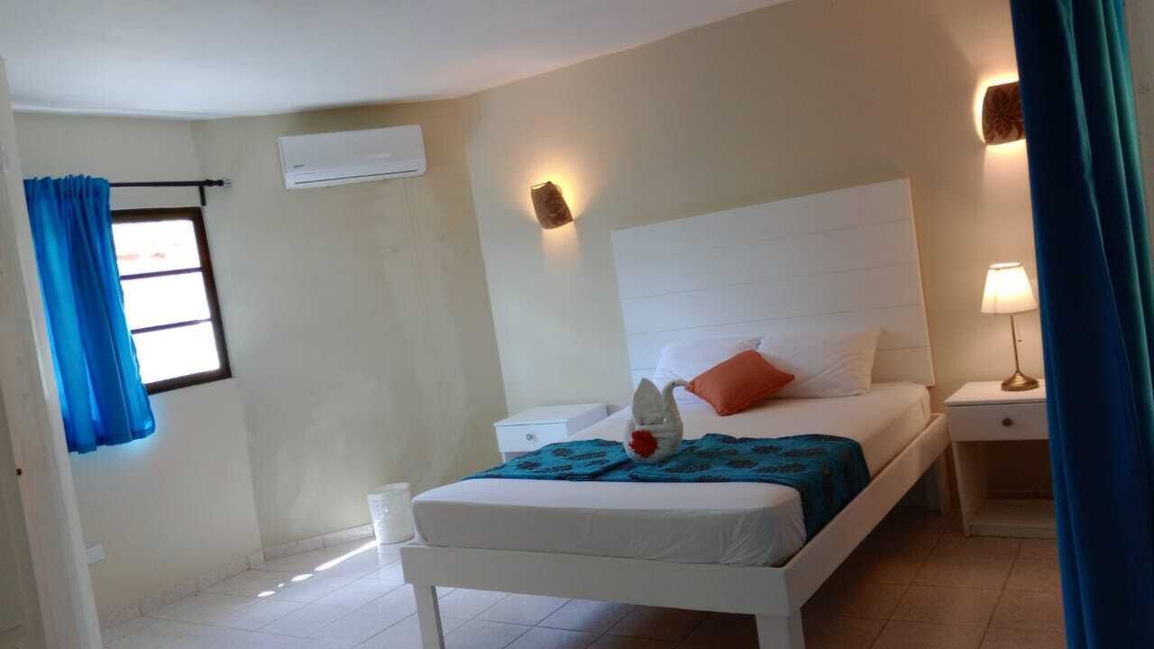 Une chambre d’hôtel avec un lit simple, une table de chevet, une lampe de chevet, un climatiseur fixé au mur et une fenêtre avec des rideaux bleus.
